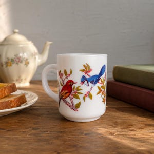 May include: A white milk glass mug featuring a colorful illustration of a red bird and a blue bird perched on flowering branches. The mug has a smooth, cylindrical body with a curved handle on the side, showcasing detailed avian artwork.