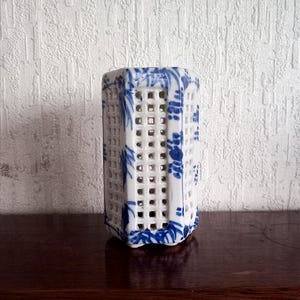 Könnte beinhalten: Eine weiße Keramikvase mit Gittermuster und blauen floralen Akzenten. Die Vase hat eine rechteckige Form mit durchbrochenen Seiten und steht auf einer dunklen Holzoberfläche.