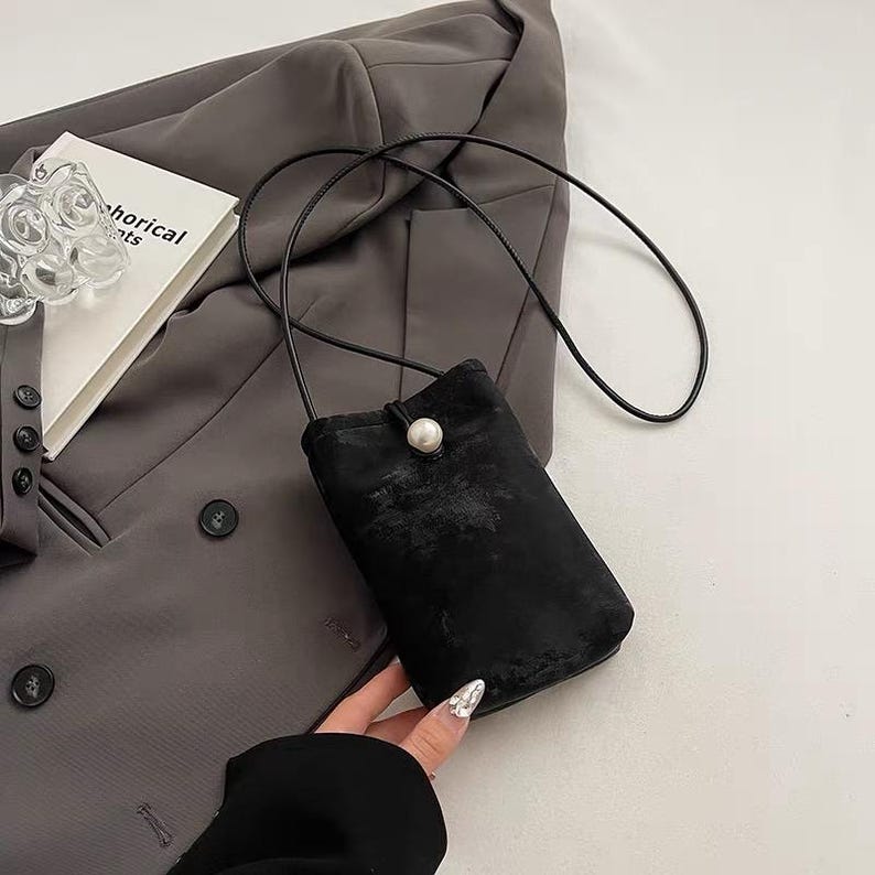 May include: A small, black velvet crossbody bag with a long, thin strap. The bag features a pearl-like button closure. The bag is held by a person's hand, and rests against a gray blazer.
