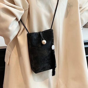 May include: A small, black crossbody bag with a long, thin strap. The bag features a pearl-like button closure and a decorative tassel. The bag is made of a soft, velvety material.
