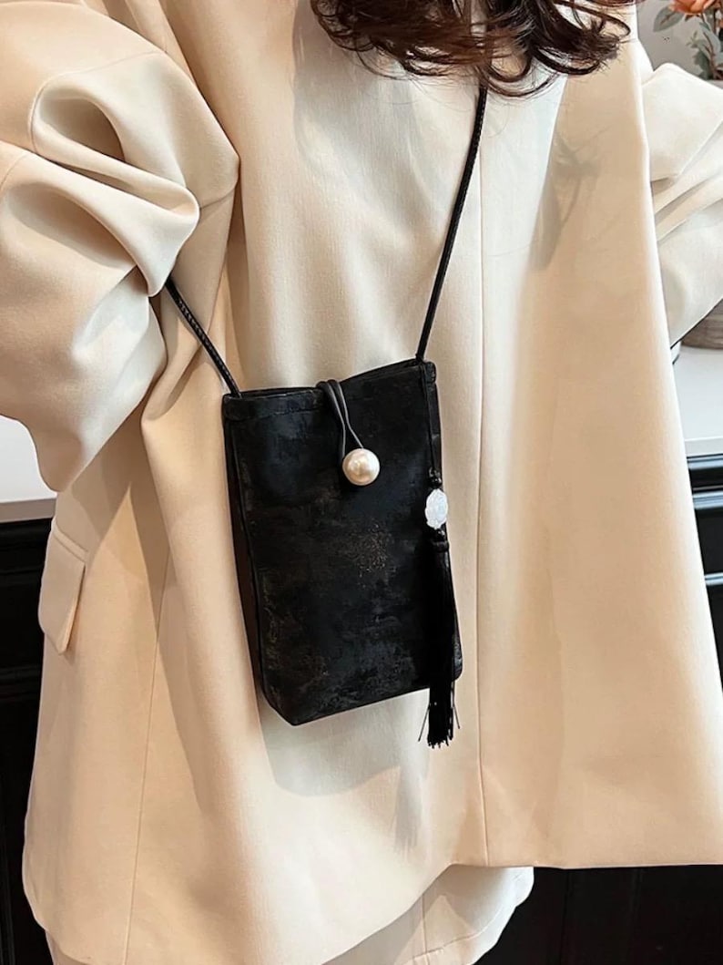 May include: A small, black crossbody bag with a long, thin strap. The bag features a pearl-like button closure and a decorative tassel. The bag is made of a soft, velvety material.