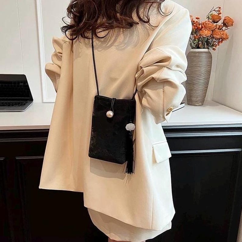 May include: A small, black velvet crossbody bag with a long, thin strap. The bag features a pearl-like bead and a decorative silver-colored embellishment. A black tassel hangs from the bottom. The bag is worn over a cream-colored blazer.