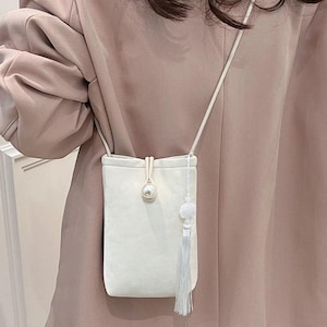 May include: A small, white, rectangular crossbody bag with a pearl accent and a tassel. The bag has a long, thin strap and a small, fluffy pom-pom detail. The bag is made of a soft, textured material.