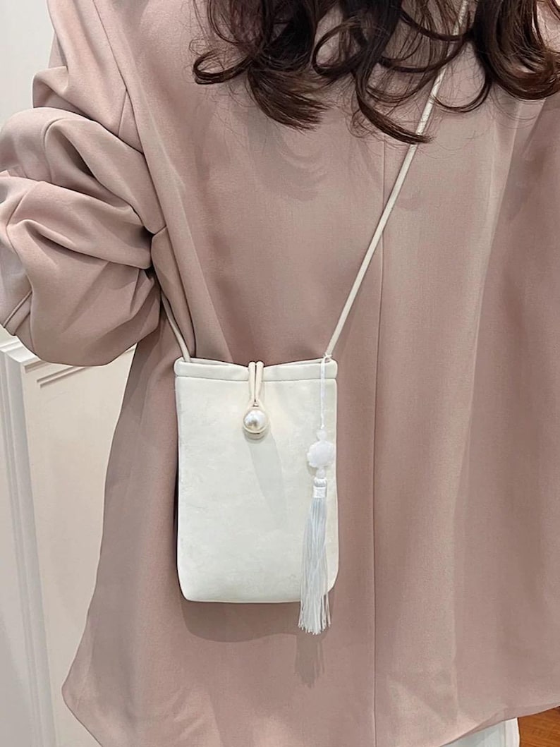 May include: A small, white, rectangular crossbody bag with a pearl accent and a tassel. The bag has a long, thin strap and a small, fluffy pom-pom detail. The bag is made of a soft, textured material.