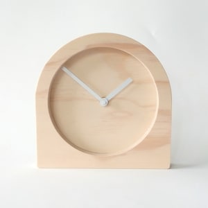 Puede incluir: Un reloj de madera con una esfera blanca y agujas blancas. El reloj tiene forma de arco y un acabado de madera clara.