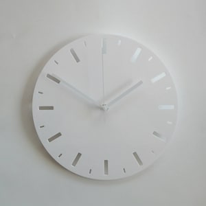 Op de afbeelding: Een witte minimalistische klok met een eenvoudig ontwerp. De wijzerplaat heeft een witte achtergrond met zwarte uurmarkeringen en een witte uur- en minutenwijzer.