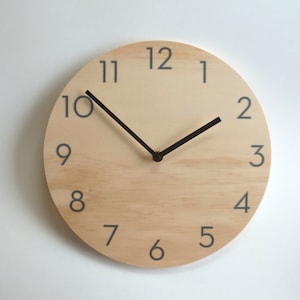 May include: A round wooden clock with black hands and grey numbers. The clock face is light wood with a darker wood grain pattern. The numbers are 1 through 12.