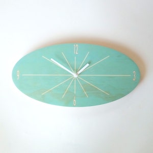 Pode incluir: Relógio turquesa oval com ponteiros brancos e números brancos. O relógio tem um design de raios de sol com linhas brancas que irradiam do centro.