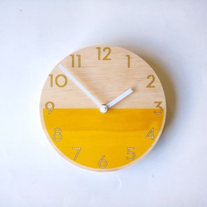 Puede incluir: Un reloj redondo de madera con manecillas blancas y una cara de madera natural y amarilla. Los números del 1 al 12 están impresos en amarillo alrededor del borde del reloj.