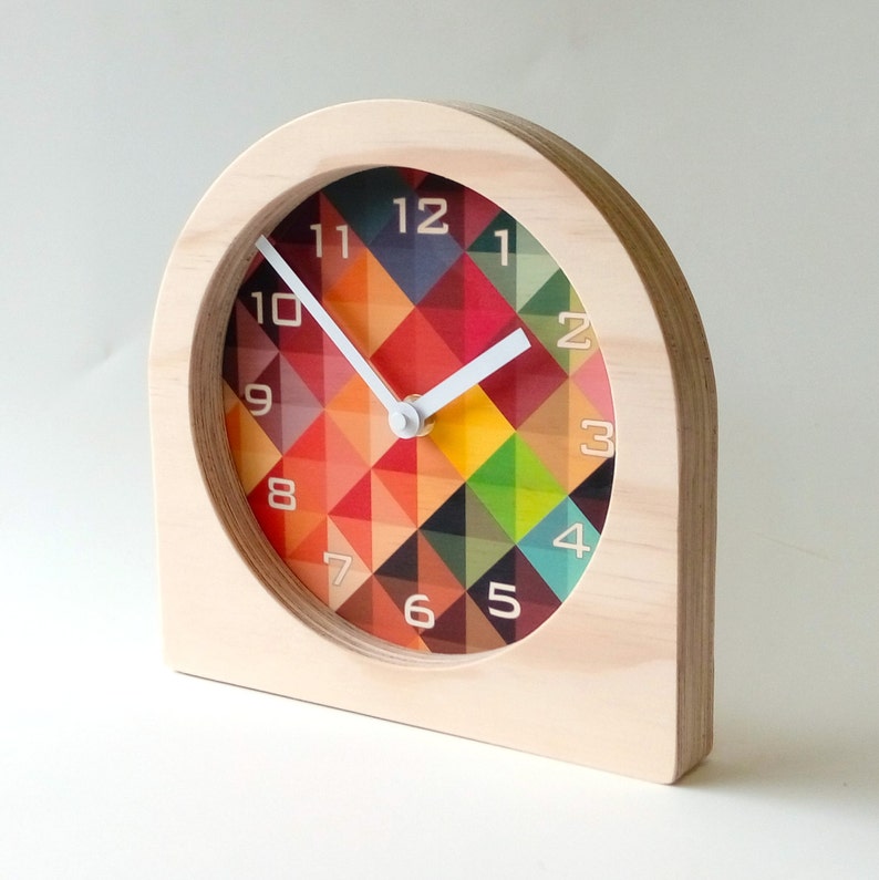 May include: A wooden desk clock with a colorful geometric design on the clock face. The clock has a light wood frame and white hands. The clock face features a pattern of red, orange, yellow, green, and blue triangles.
