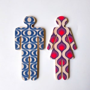 Puede incluir: Dos figuras de madera recortadas, una azul y blanca y otra morada y roja, con un patrón ondulado. Las figuras tienen forma de personas y se utilizan como letreros de baño.