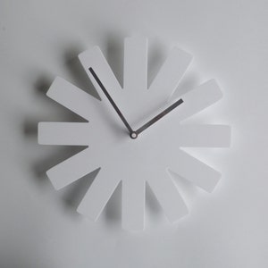 May include: White clock with a unique design featuring twelve spokes radiating from the center. The clock hands are silver.