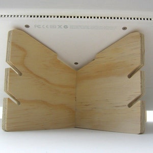 May include: A wooden laptop stand with a triangular shape and cutouts. The stand is made of light-colored wood and is designed to elevate a laptop for better ergonomics.