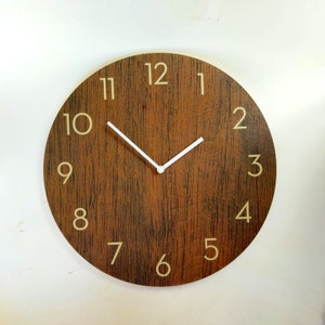 Könnte beinhalten: Eine runde Wanduhr aus Holz mit dunkelbraunem Holzmaserungsdesign. Das Zifferblatt hat beige Ziffern und weiße Zeiger. Die Uhr ist an einer weißen Wand befestigt.