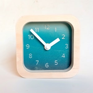 May include: A square wooden clock with a white face and black numbers. The clock face has a gradient of blue to white. The clock hands are white.