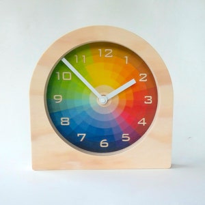 May include: A wooden clock with a rainbow coloured face and white hands. The numbers 1 through 12 are printed on the face.