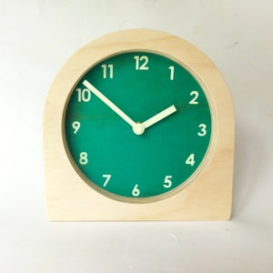 Puede incluir: Un reloj de madera con una esfera verde y números blancos. Las manecillas del reloj son blancas y el reloj está sobre una superficie blanca.