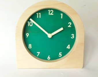 May include: A wooden clock with a green face and white numbers. The clock hands are white and the clock is sitting on a white surface.