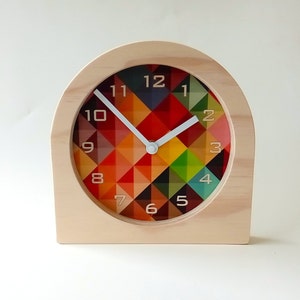 May include: A wooden desk clock with a colorful geometric design on the clock face. The clock has a light wood frame and white hands. The clock face features a pattern of red, orange, yellow, green, and blue triangles.