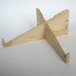 May include: A wooden laptop stand with a simple, minimalist design. The stand is made of light-colored wood and has a cross-shaped base.