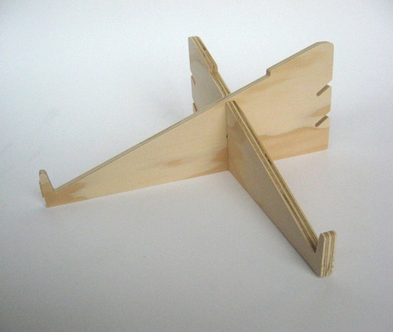 May include: A wooden laptop stand with a simple, minimalist design. The stand is made of light-colored wood and has a cross-shaped base.