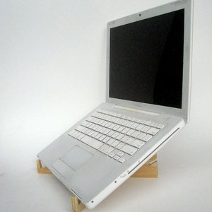 May include: A white laptop computer is elevated on a wooden stand. The laptop is closed and the screen is black. The stand is made of two pieces of wood that are joined together at a right angle.
