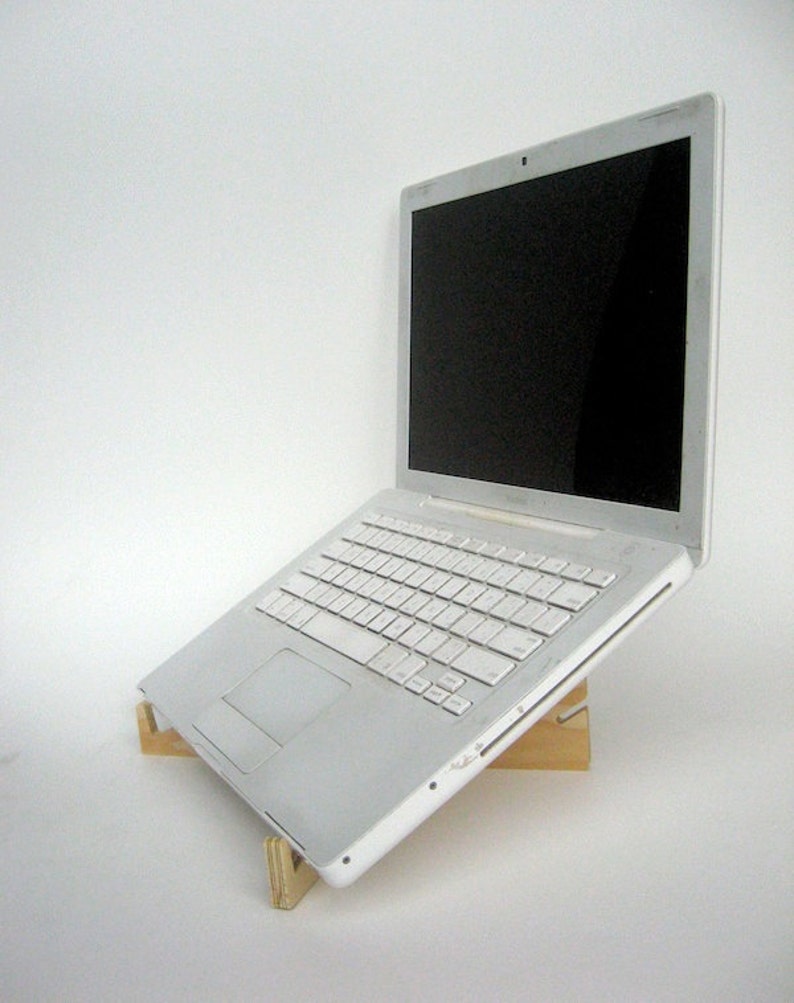 May include: A white laptop computer is elevated on a wooden stand. The laptop is closed and the screen is black. The stand is made of two pieces of wood that are joined together at a right angle.