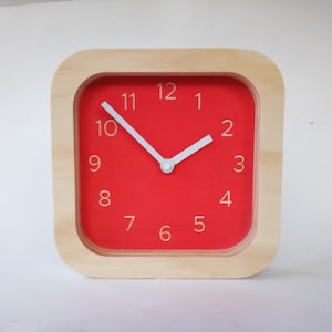 Puede incluir: Un reloj cuadrado de madera con una cara roja y manecillas blancas. Los números del 1 al 12 están impresos en blanco en la cara roja.