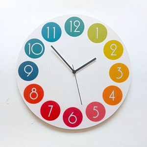 Pode incluir: Um relógio redondo colorido com números brancos em um fundo branco. O relógio tem uma mão de hora e minuto preta e uma mão de segundo branca. Os números estão em um círculo ao redor do mostrador do relógio, com o 12 no topo e o 1 à direita.