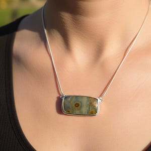 May include: A rectangular pendant necklace with a silver-colored frame. The pendant features a stone with a mix of green, brown, and gray hues. The necklace is worn on a silver chain.