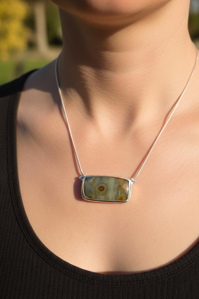 May include: A rectangular pendant necklace with a silver-colored frame. The pendant features a stone with a mix of green, brown, and gray hues. The necklace is worn on a silver chain.