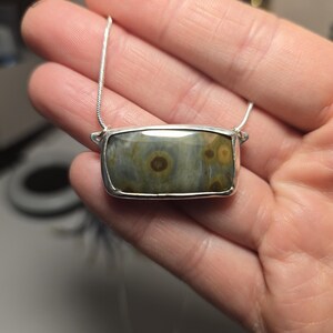 May include: A rectangular pendant necklace with a silver chain. The pendant features a polished, rectangular stone with a silver bezel. The stone has a mix of green, brown, and tan colors with circular patterns.