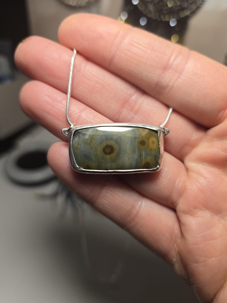 May include: A rectangular pendant necklace with a silver chain. The pendant features a polished, rectangular stone with a silver bezel. The stone has a mix of green, brown, and tan colors with circular patterns.