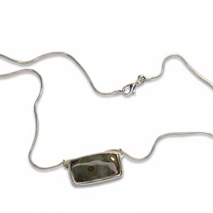 May include: A silver necklace with a rectangular pendant. The pendant features a stone with shades of green, brown, and gray. The necklace has a delicate chain and a clasp closure. The jewelry is displayed on a white background.
