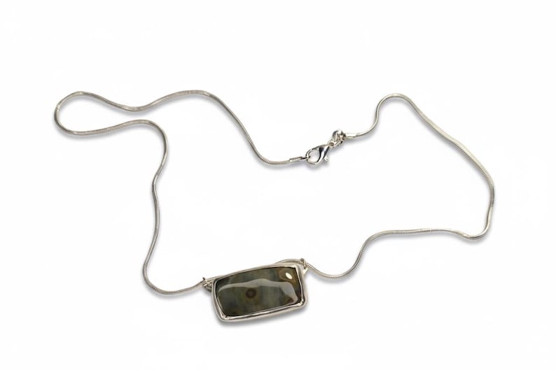 May include: A silver necklace with a rectangular pendant. The pendant features a stone with shades of green, brown, and gray. The necklace has a delicate chain and a clasp closure. The jewelry is displayed on a white background.