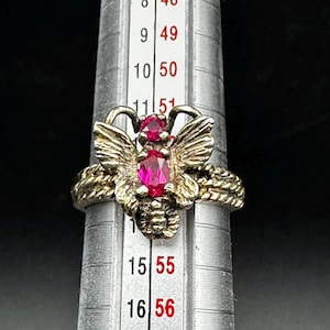 May include: A silver-toned ring featuring a butterfly design with two oval-cut pinkish-red stones. The ring is positioned on a metal ring mandrel showing size markings, with the band resting between the 11 and 12 size indicators.