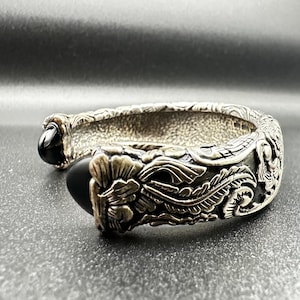 Può includere: Un bracciale a fascia color argento caratterizzato da un intricato motivo in rilievo di fiori e foglie lungo tutta la banda. Il design aperto è rifinito con due pietre ovali nere, lisce e lucide, a ciascuna estremità del bracciale.