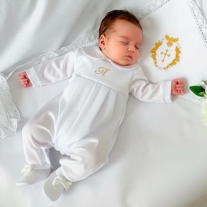 Op de afbeelding: Een slapende baby in een witte doopromper met een gouden geborduurde initiaal. De baby ligt op een wit laken naast een wit kussen met gouden borduursel en een boeket witte bloemen.