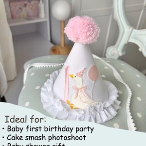 May include: White fabric party hat with a pink pom-pom, a ruffle trim, and an embroidered goose design. The hat has the number "1" and a pink balloon. A perfect accessory for a baby's first birthday.