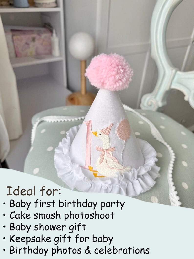 May include: White fabric party hat with a pink pom-pom, a ruffle trim, and an embroidered goose design. The hat has the number "1" and a pink balloon. A perfect accessory for a baby's first birthday.