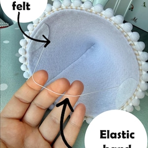 May include: A light blue felt item with a white pom-pom trim and an elastic band. The text "Soft felt" and "Elastic band" are visible. The item is held by a hand.