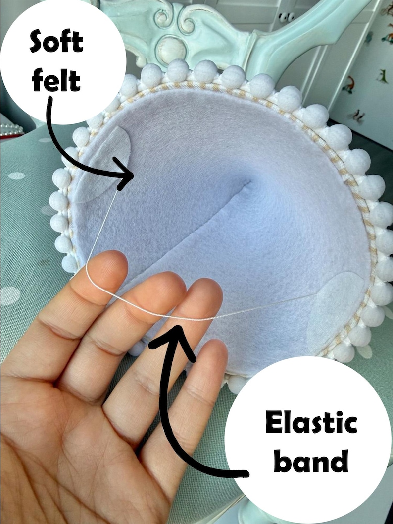May include: A light blue felt item with a white pom-pom trim and an elastic band. The text "Soft felt" and "Elastic band" are visible. The item is held by a hand.