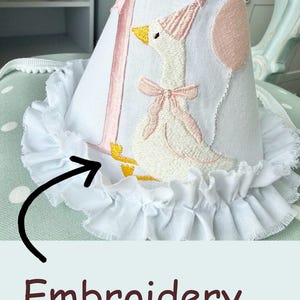May include: White fabric party hat with a pink embroidered number 1 and a goose wearing a party hat and bow. The hat has a ruffled white trim. A pink balloon is also embroidered on the hat.