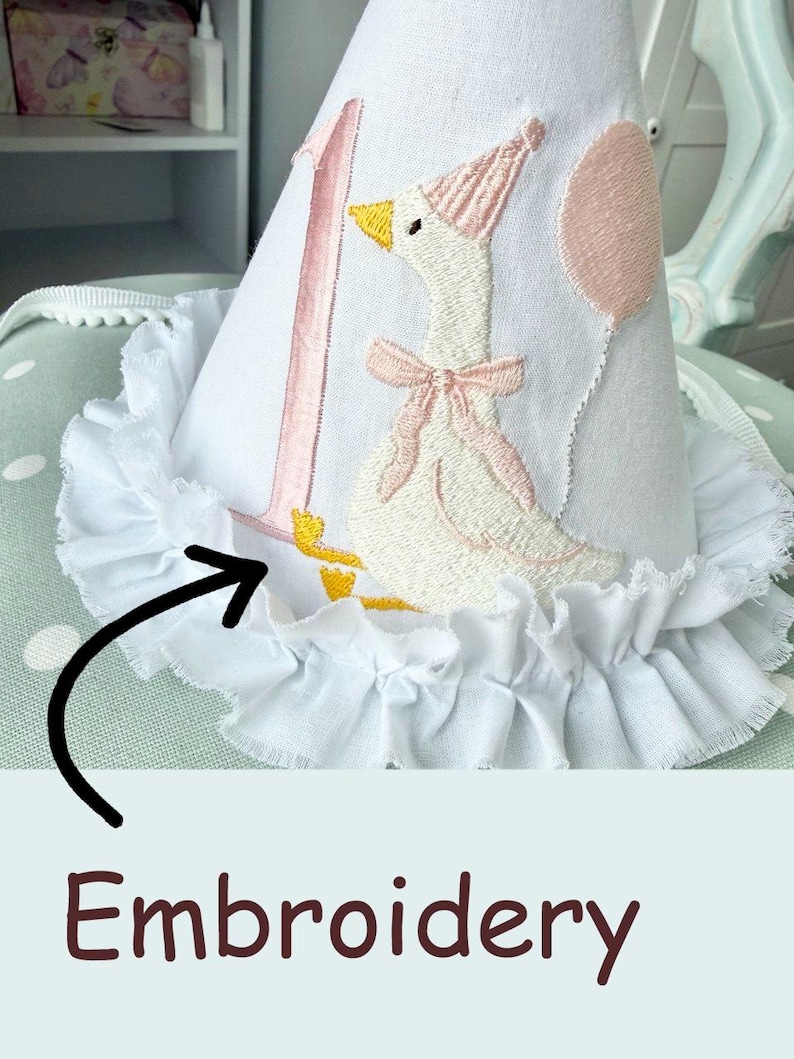 May include: White fabric party hat with a pink embroidered number 1 and a goose wearing a party hat and bow. The hat has a ruffled white trim. A pink balloon is also embroidered on the hat.