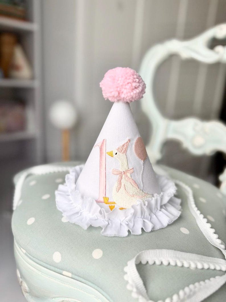 May include: White party hat with a pink pom-pom, featuring an embroidered goose with a pink bow and balloon. The number "1" is embroidered on the hat. A white ruffled trim adorns the base of the hat.