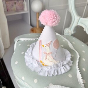 May include: A white party hat with a pink pom-pom on top. The hat features a ruffle trim and an embroidered design of a goose with a pink bow and a balloon. The number "1" is embroidered on the hat.
