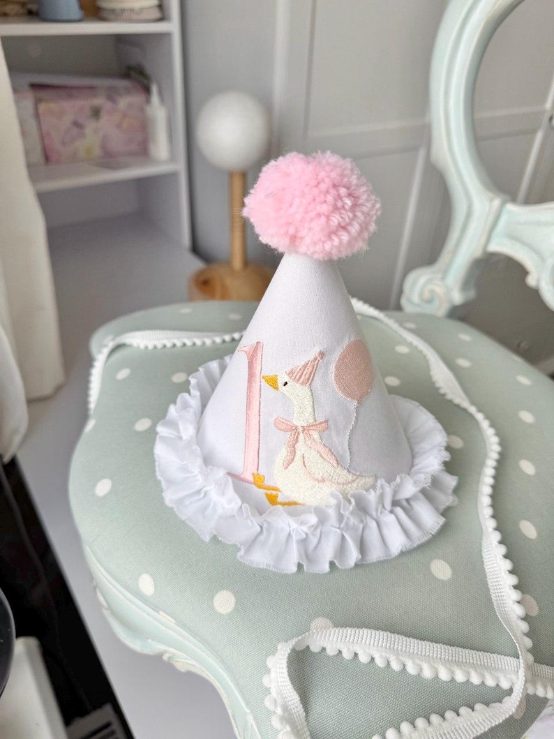 May include: A white party hat with a pink pom-pom on top. The hat features a ruffle trim and an embroidered design of a goose with a pink bow and a balloon. The number "1" is embroidered on the hat.