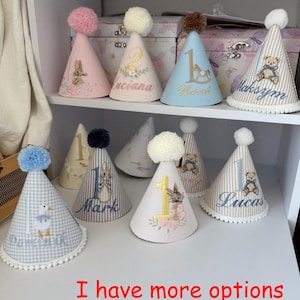 May include: A collection of personalized birthday party hats in various colors, including pink, blue, and white. Each hat features a pom-pom, a number, a name, and a decorative illustration, such as a bunny or a teddy bear. The text "I have more options in my store!" is visible.
