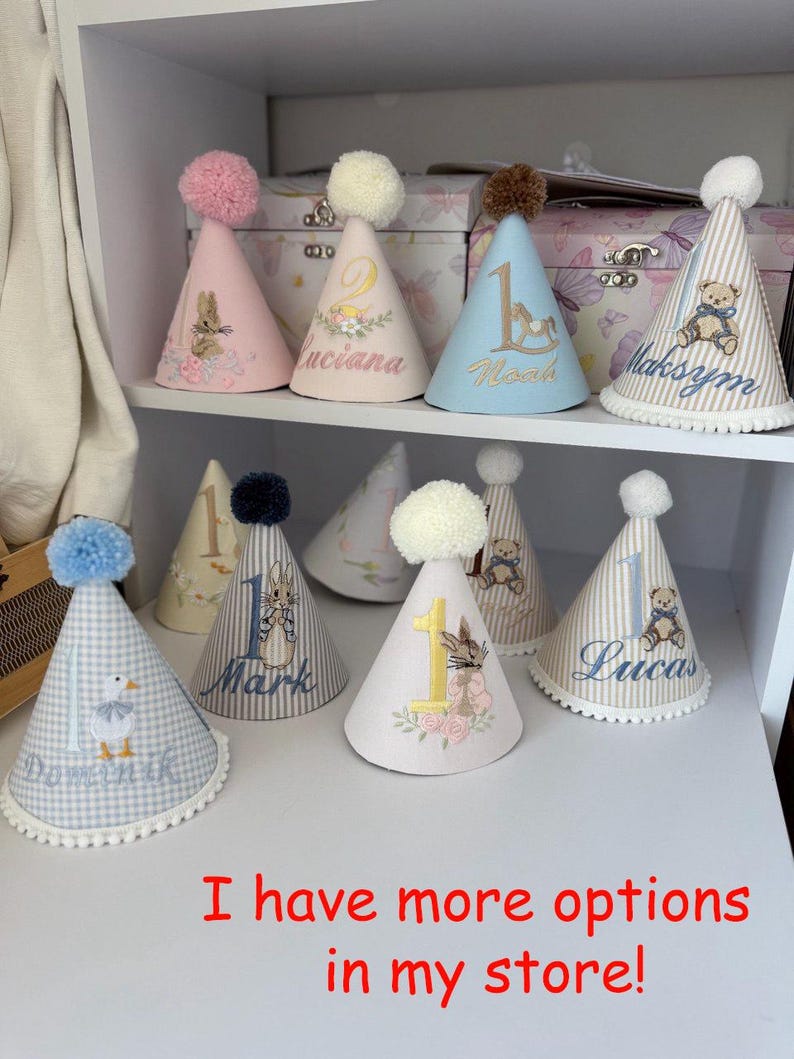 May include: A collection of personalized birthday party hats in various colors, including pink, blue, and white. Each hat features a pom-pom, a number, a name, and a decorative illustration, such as a bunny or a teddy bear. The text "I have more options in my store!" is visible.
