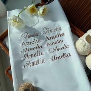 May include: White fabric with the name "Amelia" embroidered in brown script ten times. The fabric is in a wicker basket with white baby shoes and white flowers. A ball of twine is in the lower left corner.